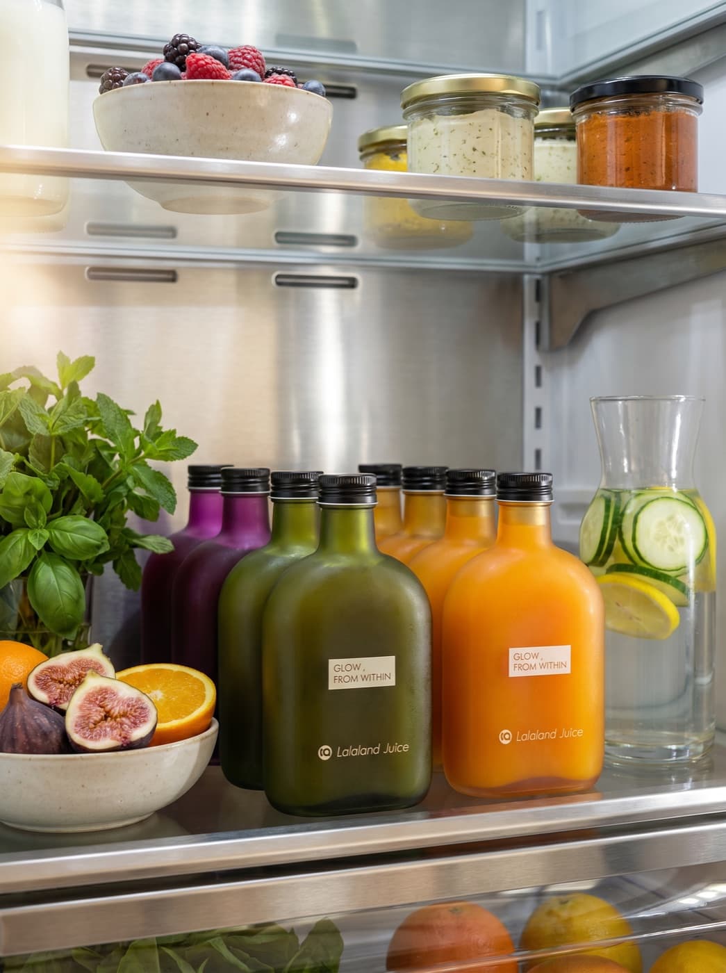 A fridge shelf lined with Lalaland juice beside fresh figs and herbs