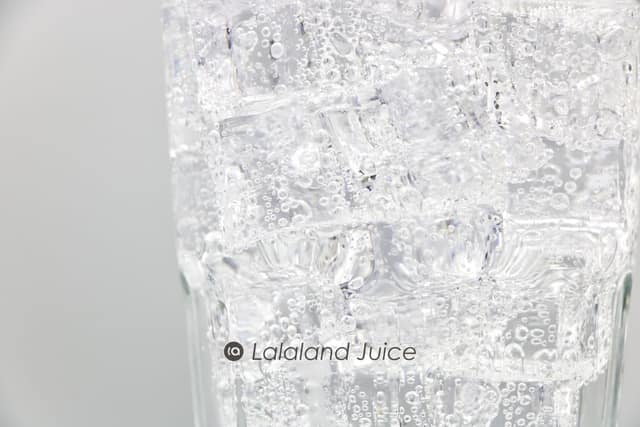 A tall glass packed with ice and sparkling water in close-up, beaded and cold — the Lalaland Juice mark set quietly within.