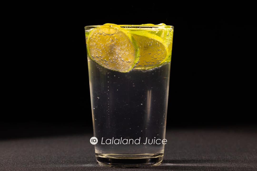 The pink Lalaland Juice flask on a marble tabletop beside a tall glass of sparkling soda water over ice, dressed with a slice of lime and loose calamansi — the fizzy, near-zero-sugar refresher.