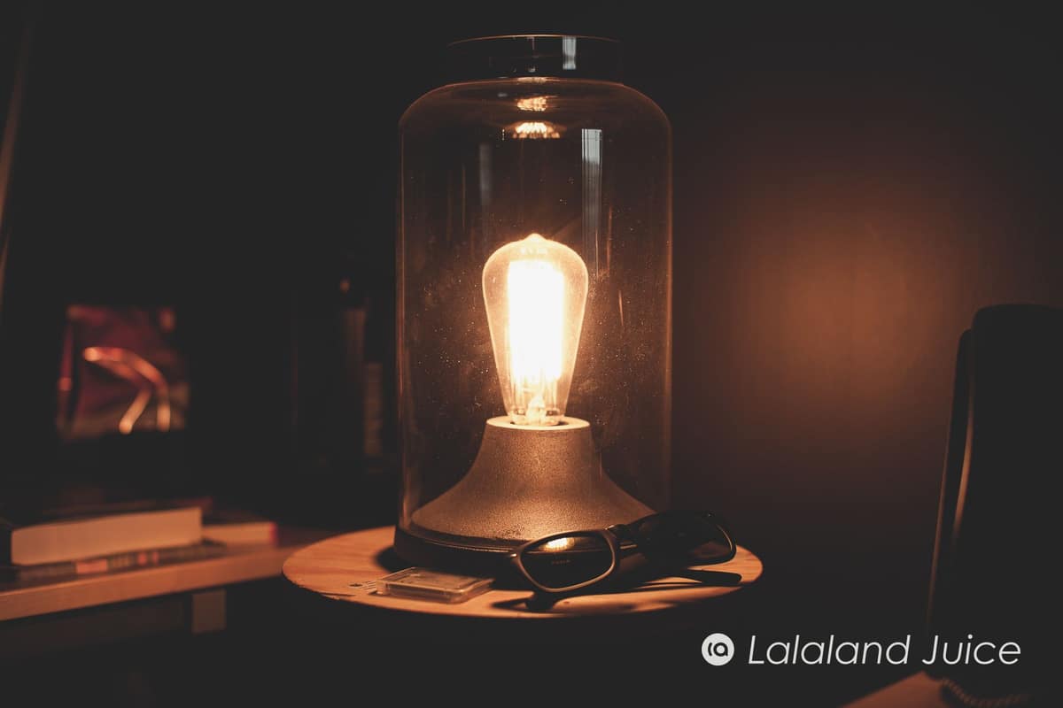 A warm Edison-bulb lamp glowing in a dimmed room at night — the dimmed room that tells your body the day is over, the Lalaland Juice mark set within.